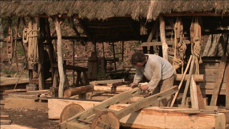 Ein Mann bearbeitet einen kleinen Wagen aus Holz mit einem Ziehmesser.