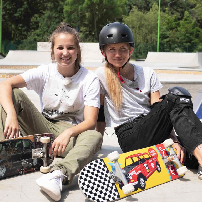 neuneinhalb-Reporterin Jana und Lea sitzen mit ihren Skateboards auf einer Skateboard-Rampe. 