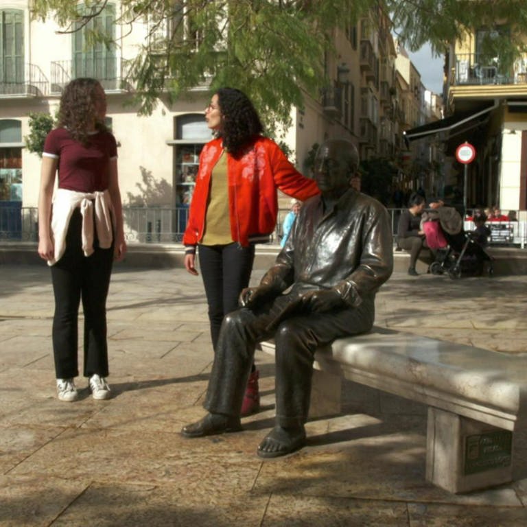 Zwei Frauen stehen auf einem Platz. Eine der beiden legt ihre Hand auf eine Bronzestatue eines Mannes, der auf einer Bank sitzt.